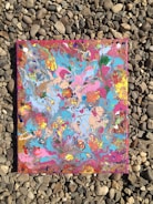 An abstract painting with vibrant swirling colors including blue, pink, orange, and yellow, placed on a background of small, smooth rocks.