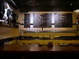 A dimly lit bar countertop with a row of beer taps in the center, beneath a menu board listing various beers available. To the left, there is a display of merchandise including shirts and a trophy, and the wall is covered in white subway tiles.