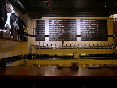A dimly lit bar countertop with a row of beer taps in the center, beneath a menu board listing various beers available. To the left, there is a display of merchandise including shirts and a trophy, and the wall is covered in white subway tiles.