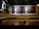 A dimly lit bar countertop with a row of beer taps in the center, beneath a menu board listing various beers available. To the left, there is a display of merchandise including shirts and a trophy, and the wall is covered in white subway tiles.