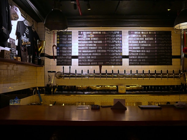 A dimly lit bar countertop with a row of beer taps in the center, beneath a menu board listing various beers available. To the left, there is a display of merchandise including shirts and a trophy, and the wall is covered in white subway tiles.