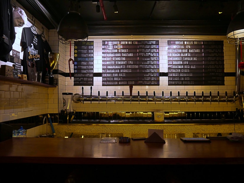A dimly lit bar countertop with a row of beer taps in the center, beneath a menu board listing various beers available. To the left, there is a display of merchandise including shirts and a trophy, and the wall is covered in white subway tiles.