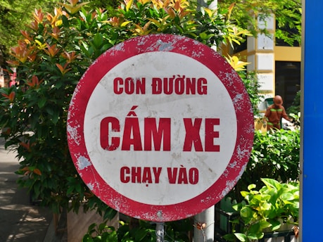 A red and white circular road sign with Vietnamese text, located in an outdoor environment surrounded by lush green foliage and a pedestrian visible in the background.