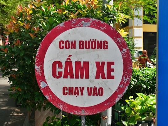 A red and white circular road sign with Vietnamese text, located in an outdoor environment surrounded by lush green foliage and a pedestrian visible in the background.