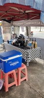A small outdoor coffee stand is set up under a red canopy with a checkered tablecloth. Various bottles, a coffee maker, and utensils are organized on the table. A large blue cooler box rests on pink stools in the foreground. Some motorcycle traffic is visible in the background.