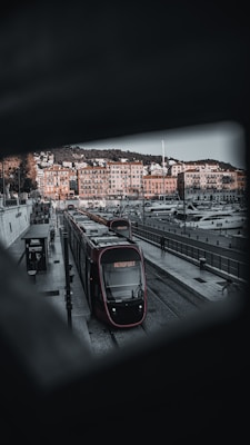 An urban scene with a red tram labeled 'Aeroport' on tracks in the foreground. The background features a line of multi-story buildings with hillside terrain behind them. The sky is overcast, and the colors are muted, giving the image a calm atmosphere. The scene is framed by dark edges, drawing focus to the tram and its surroundings.