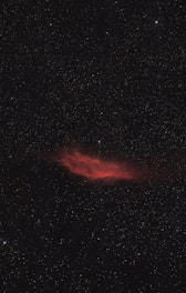 A red nebula glows amidst a dark expanse filled with countless stars. The nebula has an irregular shape and is surrounded by a shimmering field of tiny, distant stars.