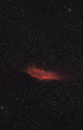 A red nebula glows amidst a dark expanse filled with countless stars. The nebula has an irregular shape and is surrounded by a shimmering field of tiny, distant stars.