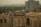 Workers are constructing a building, surrounded by scaffolding and construction materials. Some workers are wearing safety helmets and vests. The scene is urban with a partially built high-rise in the background and various trees and houses in the distance.