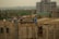 Workers are constructing a building, surrounded by scaffolding and construction materials. Some workers are wearing safety helmets and vests. The scene is urban with a partially built high-rise in the background and various trees and houses in the distance.
