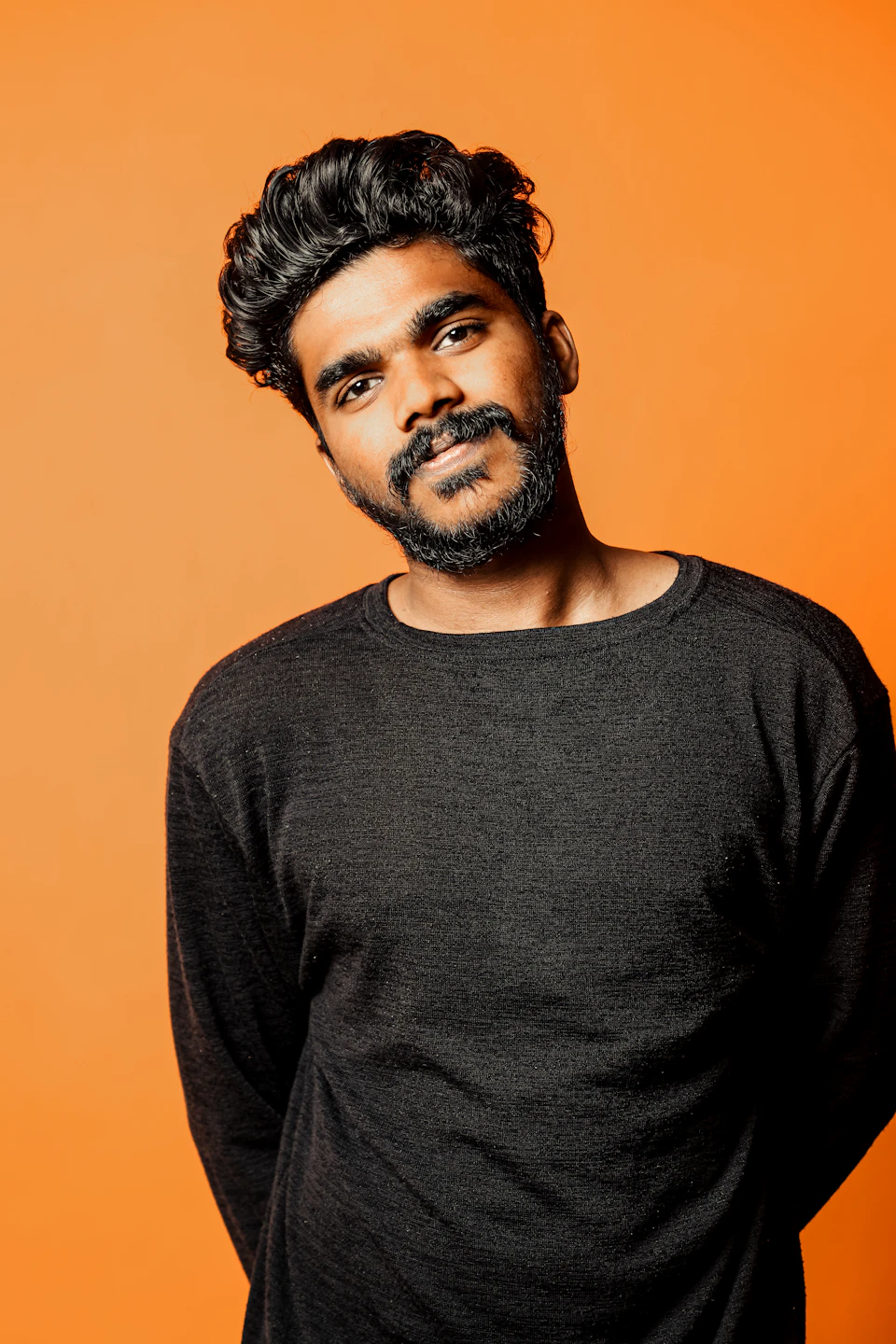 The image shows a man with dark hair and a beard wearing a black sweater, standing against an orange background. The man is looking directly at the camera with a slight smile on his face.
