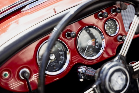 A vintage car dashboard with prominent, circular speedometers and gauges set against a glossy red background. The steering wheel is partially visible, featuring a classic design with metal spokes and a logo in the center. The intricate design and chrome finishes highlight a classic automotive aesthetic.