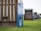 A close-up view of the back of a wooden beach chair with a blue metal plate numbered '501 W'. Several similar chairs are lined up in the background on a grassy field, each with wooden slats and striped awnings.