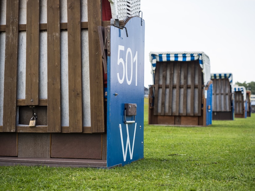 A close-up view of the back of a wooden beach chair with a blue metal plate numbered '501 W'. Several similar chairs are lined up in the background on a grassy field, each with wooden slats and striped awnings.