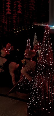 The image features festive decorations with reindeer figures covered in white and red details, surrounded by cone-shaped structures adorned with string lights. The scene is illuminated by soft, warm lighting that casts a cozy ambiance.