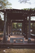 A wooden pergola structure stands in an outdoor setting, casting long shadows on the ground. The structure is complemented by concrete benches, adding a rustic appeal. A person is seen in the background near a bicycle, adding a touch of activity to the otherwise calm scene. Lush greenery surrounds the area, suggesting a park or garden setting.