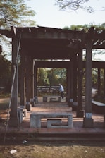 A wooden pergola structure stands in an outdoor setting, casting long shadows on the ground. The structure is complemented by concrete benches, adding a rustic appeal. A person is seen in the background near a bicycle, adding a touch of activity to the otherwise calm scene. Lush greenery surrounds the area, suggesting a park or garden setting.