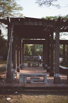 A wooden pergola structure stands in an outdoor setting, casting long shadows on the ground. The structure is complemented by concrete benches, adding a rustic appeal. A person is seen in the background near a bicycle, adding a touch of activity to the otherwise calm scene. Lush greenery surrounds the area, suggesting a park or garden setting.
