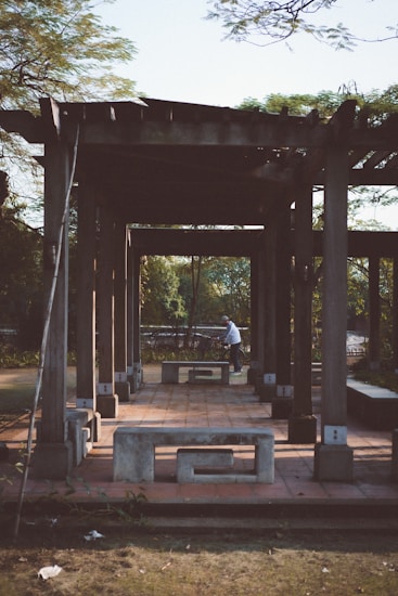 A wooden pergola structure stands in an outdoor setting, casting long shadows on the ground. The structure is complemented by concrete benches, adding a rustic appeal. A person is seen in the background near a bicycle, adding a touch of activity to the otherwise calm scene. Lush greenery surrounds the area, suggesting a park or garden setting.