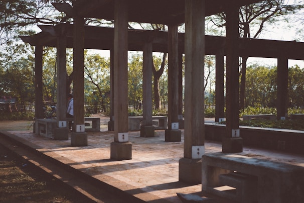 A wooden pavilion structure with multiple columns casting shadows on the ground. There are benches made of concrete blocks inside the pavilion. Surrounding the structure are trees with green foliage, indicating a natural, outdoor park setting. Sunlight filters through the trees, creating a serene atmosphere.