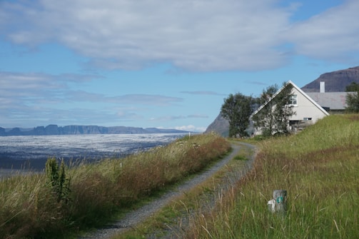 A picturesque landscape featuring a gravel path leading towards a white house with a slanted roof surrounded by tall grass. In the background, a vast expanse of dramatic mountains meets a glacier under a partly cloudy sky. Trees are scattered around the house, adding to the serene and remote ambiance.