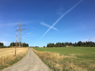 A rural landscape featuring a dirt path flanked by grass fields, with a tall metal electricity pylon on the left. The sky is clear, displaying a deep blue color with white contrails stretching across. A line of trees in the distance separates the fields from the horizon.