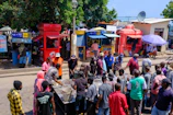 A bustling street scene in an urban setting with a group of people gathered around a table or stall. The backdrop includes a variety of brightly colored kiosks, likely for mobile network services, with some individuals engaging with vendors. Trees and a blue sky are visible in the background, adding a natural element to the busy marketplace atmosphere.