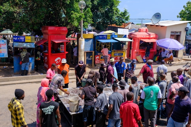 A bustling street scene in an urban setting with a group of people gathered around a table or stall. The backdrop includes a variety of brightly colored kiosks, likely for mobile network services, with some individuals engaging with vendors. Trees and a blue sky are visible in the background, adding a natural element to the busy marketplace atmosphere.