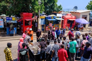 A bustling street scene in an urban setting with a group of people gathered around a table or stall. The backdrop includes a variety of brightly colored kiosks, likely for mobile network services, with some individuals engaging with vendors. Trees and a blue sky are visible in the background, adding a natural element to the busy marketplace atmosphere.