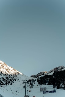 A snowy ski resort featuring a chairlift leading up a mountain with scattered skiers below. The slopes are bordered by rocky peaks and dense evergreen trees. Bright sunlight casts shadows across the snow, while a large Samsung advertisement is visible in the foreground.