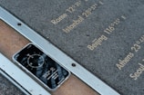 A smartphone rests face-up on a metal and stone surface displaying a compass app with the reading 352 degrees North. Etched inscriptions on the stone list various city names and their longitudinal coordinates, including Rome, Istanbul, Beijing, Athens, and Seoul.
