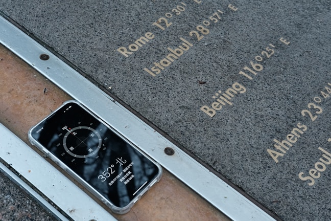 A smartphone rests face-up on a metal and stone surface displaying a compass app with the reading 352 degrees North. Etched inscriptions on the stone list various city names and their longitudinal coordinates, including Rome, Istanbul, Beijing, Athens, and Seoul.