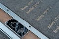 A smartphone rests face-up on a metal and stone surface displaying a compass app with the reading 352 degrees North. Etched inscriptions on the stone list various city names and their longitudinal coordinates, including Rome, Istanbul, Beijing, Athens, and Seoul.