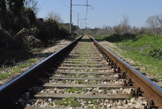 A straight railway track extends into the distance, flanked by lush greenery and shrubs on either side. The track is made of metal rails and wooden sleepers, with gravel filling the gaps. Overhead, clear skies are visible, and the surrounding vegetation hints at a rural or countryside setting.