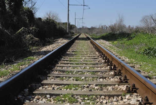 A straight railway track extends into the distance, flanked by lush greenery and shrubs on either side. The track is made of metal rails and wooden sleepers, with gravel filling the gaps. Overhead, clear skies are visible, and the surrounding vegetation hints at a rural or countryside setting.