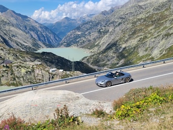 A scenic mountain landscape featuring a winding road running alongside a turquoise lake. A sleek, silver convertible sports car is driving along the road with a person inside, under a bright blue sky filled with fluffy clouds. The scene is surrounded by rocky, green hillsides.