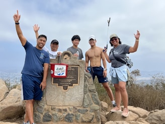 Five people are posing at a mountain viewpoint next to a stone plaque. They are smiling and making celebratory gestures. The group includes both men and women, with one person holding a trash picker and a bucket labeled 'Clean Trails.' The background reveals a cloudy sky and a scenic view.