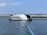A modern barge with a sleek design is stationed on the water, featuring a structure labeled 'The Ocean Cleanup.' It has a serial number '007' and is equipped with an extended boom-like apparatus floating on the water's surface. The backdrop includes a rocky breakwater with a few people walking along it and a clear, blue sky.