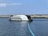 A modern barge with a sleek design is stationed on the water, featuring a structure labeled 'The Ocean Cleanup.' It has a serial number '007' and is equipped with an extended boom-like apparatus floating on the water's surface. The backdrop includes a rocky breakwater with a few people walking along it and a clear, blue sky.