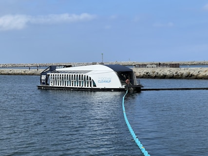 A modern barge with a sleek design is stationed on the water, featuring a structure labeled 'The Ocean Cleanup.' It has a serial number '007' and is equipped with an extended boom-like apparatus floating on the water's surface. The backdrop includes a rocky breakwater with a few people walking along it and a clear, blue sky.
