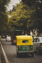A yellow and green auto rickshaw is driving down a tree-lined street alongside other vehicles. The road is bordered by lush green trees and the scene suggests a typical city traffic environment.