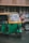 A green and yellow auto rickshaw is parked on a street next to a brown car. The auto rickshaw has a license plate with the number 'KA 03 AA 5184'. The background features a building with a patterned facade and weathered walls.