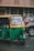 A green and yellow auto rickshaw is parked on a street next to a brown car. The auto rickshaw has a license plate with the number 'KA 03 AA 5184'. The background features a building with a patterned facade and weathered walls.