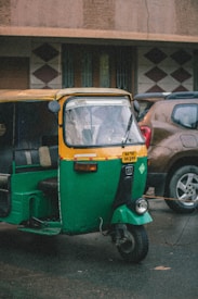 A green and yellow auto rickshaw is parked on a street next to a brown car. The auto rickshaw has a license plate with the number 'KA 03 AA 5184'. The background features a building with a patterned facade and weathered walls.