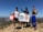 Three people are standing on a rocky hilltop under a clear blue sky. They are holding a banner with 'Clean Trails' written on it and a bucket labeled 'Clean Up.' The individuals are dressed in athletic clothing, suggesting they are participating in an outdoor event, likely related to environmental conservation.