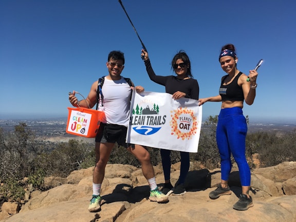 Three people are standing on a rocky hilltop under a clear blue sky. They are holding a banner with 'Clean Trails' written on it and a bucket labeled 'Clean Up.' The individuals are dressed in athletic clothing, suggesting they are participating in an outdoor event, likely related to environmental conservation.