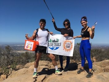 Three people are standing on a rocky hilltop under a clear blue sky. They are holding a banner with 'Clean Trails' written on it and a bucket labeled 'Clean Up.' The individuals are dressed in athletic clothing, suggesting they are participating in an outdoor event, likely related to environmental conservation.