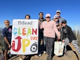 A group of people is standing outdoors holding a 'National Clean Up Day' banner. They appear to be on a mountain or elevated terrain, with a clear blue sky in the background. Some of them are holding cleaning equipment, such as a bucket and a grabber tool. They are dressed in casual activewear and are smiling at the camera.