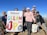A group of people is standing outdoors holding a 'National Clean Up Day' banner. They appear to be on a mountain or elevated terrain, with a clear blue sky in the background. Some of them are holding cleaning equipment, such as a bucket and a grabber tool. They are dressed in casual activewear and are smiling at the camera.