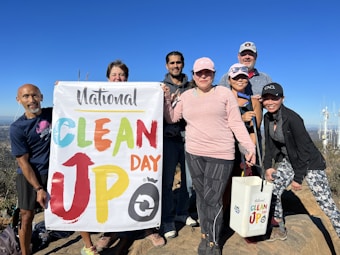 A group of people is standing outdoors holding a 'National Clean Up Day' banner. They appear to be on a mountain or elevated terrain, with a clear blue sky in the background. Some of them are holding cleaning equipment, such as a bucket and a grabber tool. They are dressed in casual activewear and are smiling at the camera.